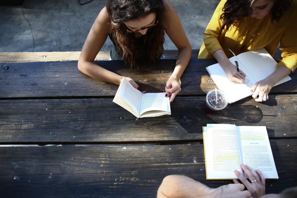 Students sit at a wooden table on the street and read