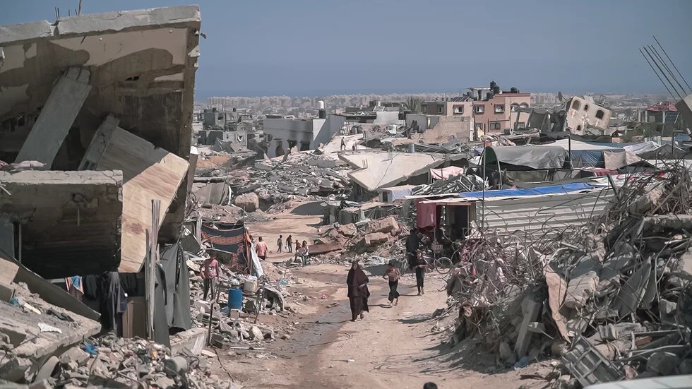 Foto: Zerstörte Gebäude und Trümmer in Gaza, mehrere Personen gehen eine staubige Straße entlang.